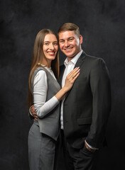 Studio portrait of smiling man in classic suit and friendly woman in trendy pantsuit. Dark background. Concept of teamwork, confidence, and positive business attitude