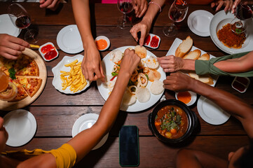 Close-up of friends sharing a delicious meal and drinks at a lively dinner party, featuring seafood, fries, and wine on a wooden table