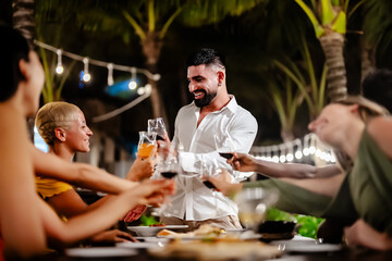 Group of diverse friends raising glasses in a joyful toast at a tropical outdoor party, celebrating together under palm trees and string lights