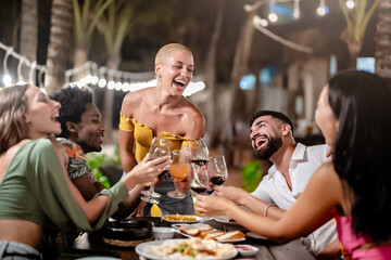 Group of diverse friends raising glasses in a joyful toast at a tropical outdoor party, celebrating together under palm trees and string lights
