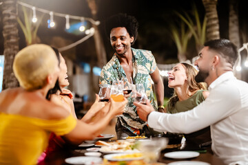 Group of diverse friends raising glasses in a joyful toast at a tropical outdoor party, celebrating together under palm trees and string lights