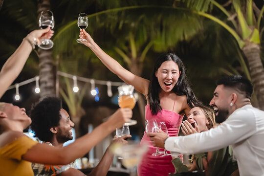 Group of diverse friends raising glasses in a joyful toast at a tropical outdoor party, celebrating together under palm trees and string lights
