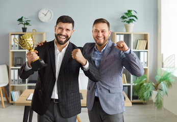 Happy young business people holding the trophy award for success in business standing in office together. Male colleagues rejoicing in achievements at work receiving a cup for best job reward.
