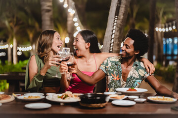 Group of happy friends sharing laughs and drinks at an outdoor dinner party under palm trees and string lights, enjoying food and festive vibes during a tropical night celebration