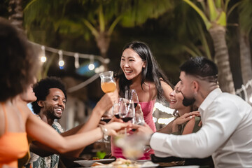 Group of happy friends sharing laughs and drinks at an outdoor dinner party under palm trees and string lights, enjoying food and festive vibes during a tropical night celebration
