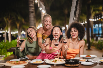 Group of joyful women toasting with wine glasses at an outdoor tropical party at night, smiling and celebrating together under string lights surrounded by palm trees and a festive atmosphere
