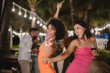 Two joyful women dancing with wine glasses during a fun tropical night party, surrounded by palm trees and string lights, celebrating friendship and enjoying the beach nightlife