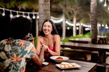 A couple toasting red wine over a romantic dinner at a tropical outdoor restaurant, featuring pizza, fries, and warm lighting for a cozy, intimate dining experience