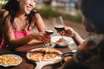 Close-up of a couple toasting red wine over a romantic dinner at a tropical outdoor restaurant, featuring pizza, fries, and warm lighting for a cozy, intimate dining experience