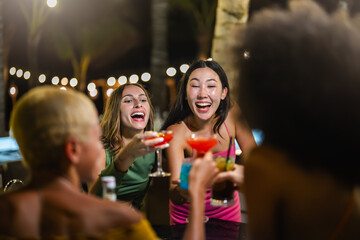 Group of young women toasting colorful cocktails and laughing together during a fun outdoor night...