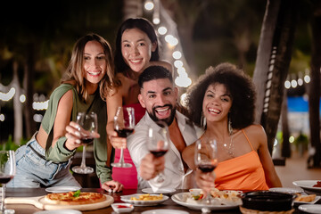 Group of friends toasting with red wine during an outdoor dinner party at night, smiling and enjoying a festive meal under string lights