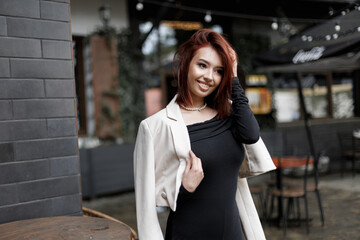 Smiling young woman with red hair wearing an elegant off-shoulder black dress, standing outdoors near a cafe. Urban street style, confidence, and charm in a casual city setting. Beautiful happy lady