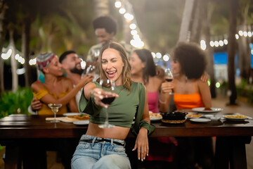 Joyful woman raising her glass at a festive outdoor dinner party with a diverse group of friends, celebrating and laughing under string lights in a tropical evening setting