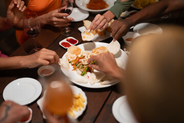Close-up of friends sharing a delicious meal and drinks at a lively dinner party, featuring seafood, fries, and wine on a wooden table
