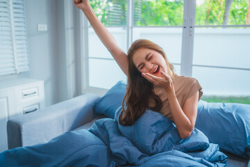 Asian housewife wakes up in the morning, stretching happily on her bed with a blue blanket. Natural sunlight fills the room, creating a fresh and comfortable atmosphere by the window.