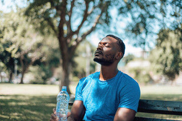 A man in his thirties relaxes on a park bench, enjoying a moment of peace under the sun while holding a bottle of water. He is coping with the intense summer heatwave