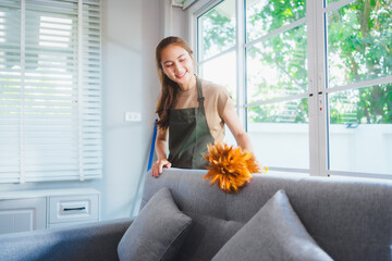 Asian housewife wearing an apron is cleaning the sofa beside a large window. Sunlight streams in,...