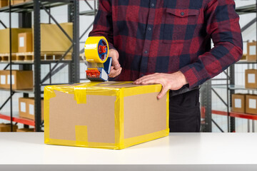 Box in packer hands. Worker seals box with tape. Cropped packer at work. Parcel packing in warehouse. Storekeeper with tape dispenser in hand. Packer hands at work. Parcel fulfillment