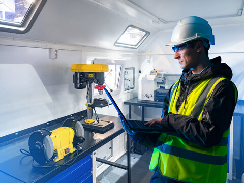 Engineer in safety gear using a laptop in a mobile workshop with industrial tools and machines, focusing on technical diagnostics and machinery setup. – Industrial Engineering.