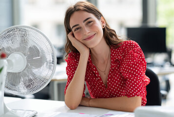 In a vibrant office, a woman relaxes at her desk, smiling as a fan cools her. The bright interior contrasts with the extreme summer heat outside, capturing a carefree moment