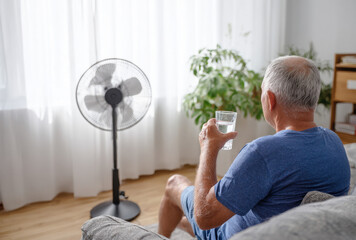 An elderly man sits comfortably on a sofa, holding a glass of water as he seeks relief from the oppressive summer heatwave. A fan blows cool air nearby, offering solace