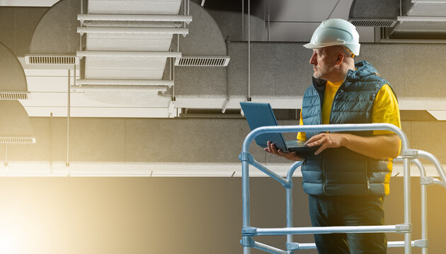 Technician with helmet and laptop inspects ceiling ventilation system from lift platform in modern industrial building. - Ventilation Installation.
