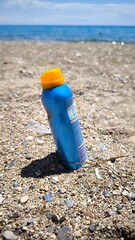 A blue sunscreen spray bottle stuck in pebbly sand near the sea