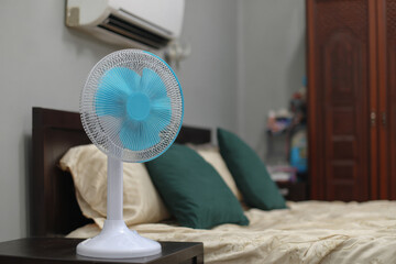 A blue and white standing fan offers relief from the intense summer heat wave in a tranquil bedroom setting with soft bedding and green pillows