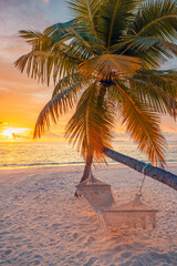 Closeup sunset view palm tree with hammock swing over golden sand sea. Romantic inspire carefree tropical landscape with vivid sky, perfect for summer vacation, relaxation, and dreamy beach background
