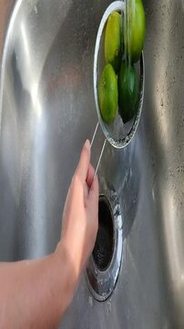 Unas manos de mujer lavando varios limones verdes dentro de un colador de metal con agua, en un fregadero en la cocina. Video vertical
