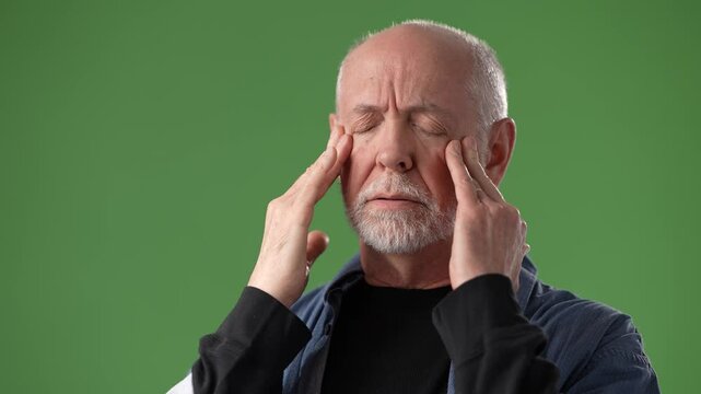 A middle-aged man with gray hair appears to be experiencing stress, holding his temples in a studio setup with a green screen background.