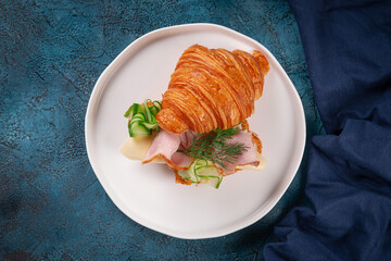 Fresh buttery croissant filled with thinly sliced ham, creamy cheese, cucumber ribbons and fresh dill. Served on a white plate over blue stone background.