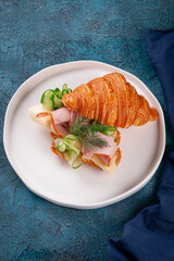 Fresh buttery croissant filled with thinly sliced ham, creamy cheese, cucumber ribbons and fresh dill. Served on a white plate over blue stone background.