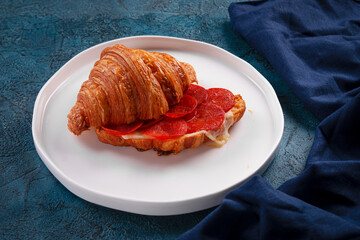 Appetizing croissant sandwich filled with melted cheese and spicy pepperoni slices. Served on a white plate with elegant dark blue napkin on textured background