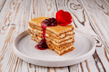 Close-up of honey layer cake with creamy filling, topped with berry jam and decorated with a fresh red rose. Served on a white ceramic plate, ideal for dessert menu or pastry showcase