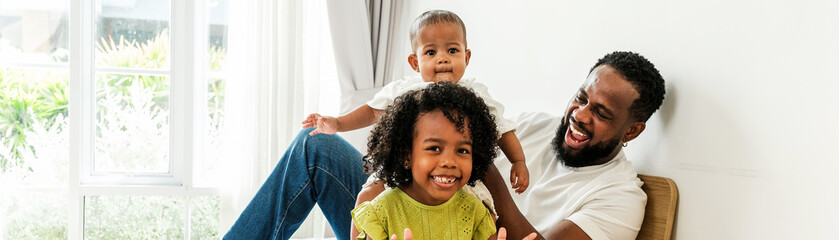 Joyful Black father plays with his baby and daughter on a bright bed in a cozy home, celebrating diverse family love and Father’s Day. A candid moment full of laughter, bonding, and parenthood joy.