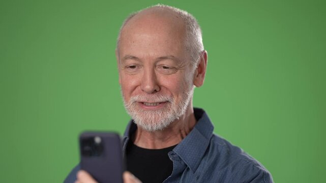 A mature man smiles and waves while using a smartphone, set against a bright green screen. He appears cheerful and connected, enjoying a virtual conversation.