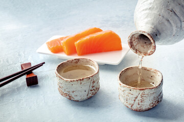 Sake poured into ochoko, traditional cup, with salmon sashimi in the background and chopsticks. Traditional Japanese food and drink