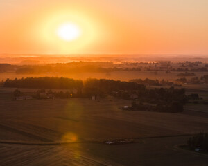 View from a hot air balloon at sunset, the sun over fields and forests, fog and haze in the distance. The yellow-brown tones of the golden hour.