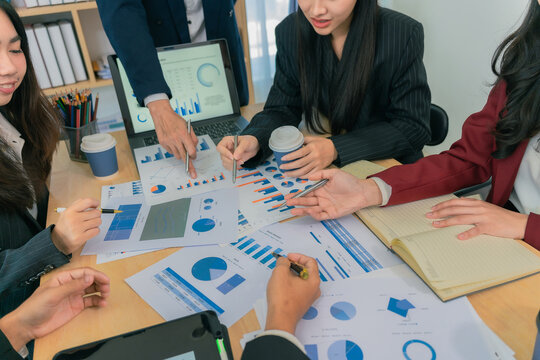 Group of young Asian businesswomen presenting financial sales report to colleagues in a diverse team, pointing at tablet screen on electronic board.