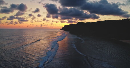 Capturing breathtaking aerial views of a sunset over the ocean, illuminating crashing waves against Bali's dramatic cliffs and highlighting the warm hues of the sky