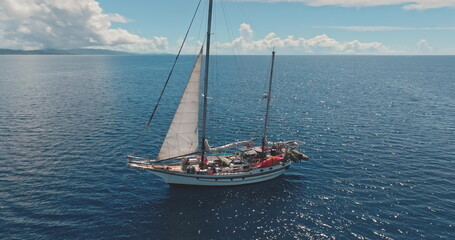 Aerial drone footage of a ketch sailing near a tropical island on a sunny day, with white sails and a wooden deck, surrounded by blue waters and a distant coastline. Wild sea nature travel background