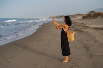 Brunette Woman in Black Dress Taking Photos on the Beach with Straw Bag