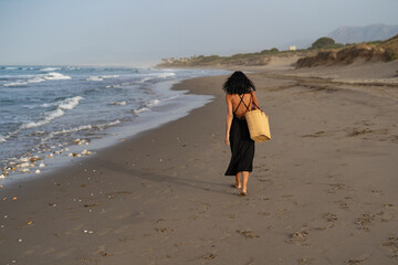 Brunette Woman in Black Dress Walking on the Beach with Straw Bag