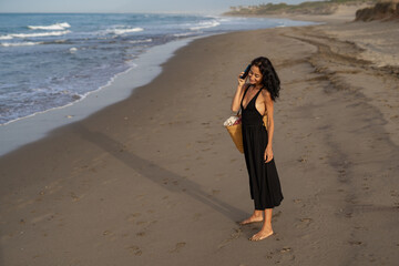 Brunette Woman in Black Dress Talking on Phone at the Beach with Straw Bag