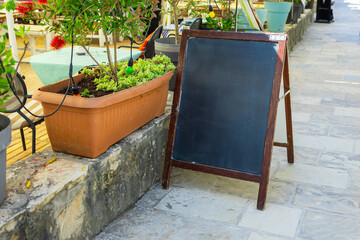 Blank wooden chalkboard stands beside flower planter on stone walkway. Outdoor signage, seasonal menu display, and cozy cafe atmosphere. Copy space
