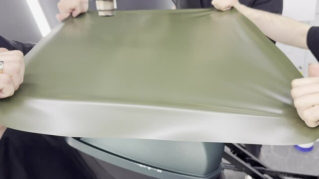 A specialist in wrapping a car with green khaki colored vinyl or ppf film in the process of work. Car wrapping specialists cover the car with green vinyl film. Khaki green vinyl foil. Selective focus.