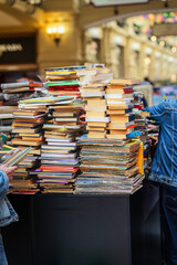 Stacked used books, sale on market. Messy stacks of old and new books on market table with people browsing. Concept of books, used literature, vintage finds and casual book market