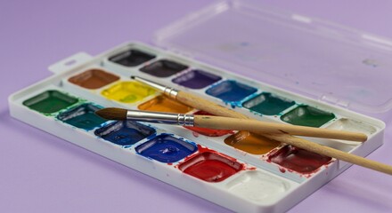A watercolor palette with vibrant paints and two paintbrushes lying across it on a soft purple surface.