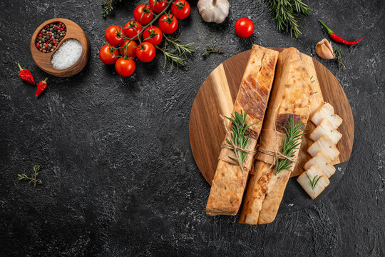 Sliced cured pork fatback with herbs, tomatoes, spices, and garlic arranged on a wooden board over dark stone background top view. copy space